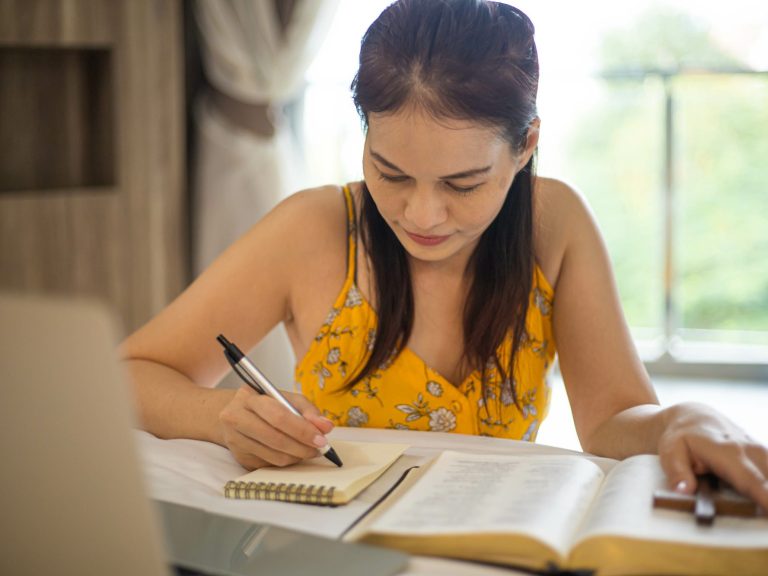 Une femme écrit dans un carnet, entourée de documents, dans un environnement lumineux.