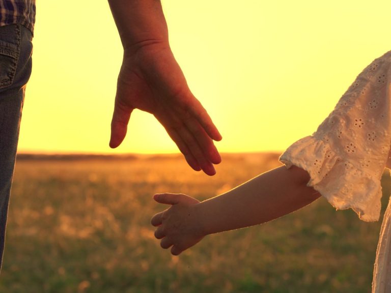 Main adulte tenant la main d'un enfant, silhouette contre un coucher de soleil.
